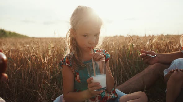 A Beautiful Preschool Girl Drinks Milk Through a Straw and Talking with Her Family at a Picnic in a