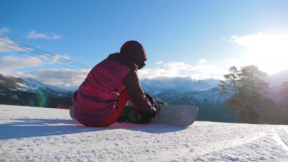 Girl with Snowboard Sitting on Top and Enjoying Mountain Landscape. Sport Woman in Snowy Mountains