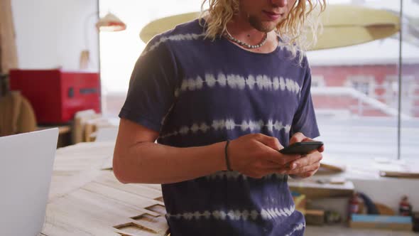 Caucasian male surfboard maker standing in his studio and using his smartphone