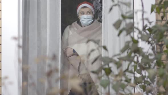 Ill Young Man in Covid19 Face Mask Wrapped in Blanket Looking Out the Window on New Year's Eve