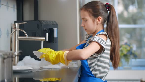 Side View of Schoolgirl in Apron and Gloves Washing Dishes After Lunch