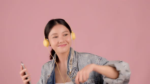 Happy pretty Asian teenager girl dancing and listening music by smartphone on pink background.