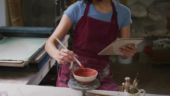 Female caucasian potter wearing face mask holding digital tablet painting pot on potters wheel at po