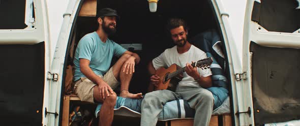 Two friends sitting in back of a van having fun and playing the guitar