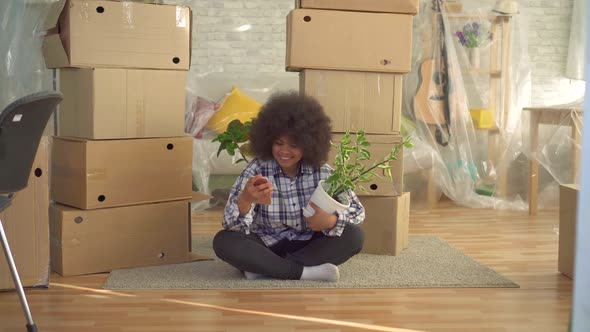 African Woman with an Afro Hairstyle with a Plant in His Hand Uses the Phone Sitting on the Floor