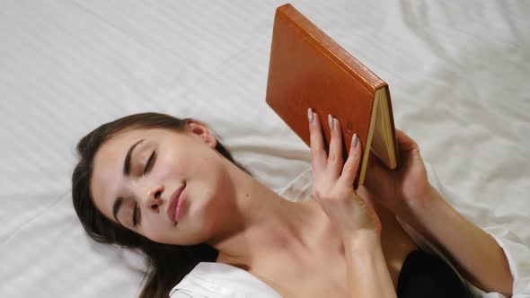 Young Pretty Woman Lying on Bed in Her Apartament with a Closed Notebook in Hand