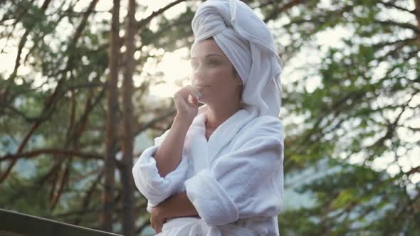 Young Woman in a Bathrobe and Towel on the Terrace on a Background of Mountains and Forest