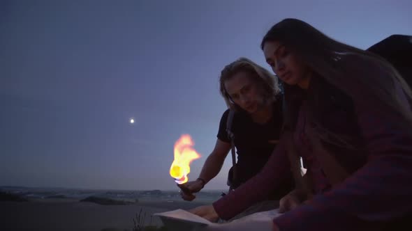 Young Couple Using a Flaming Torch To Read a Map at Dusk in the Wilderness During a Hiking Trip with