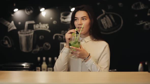 A Beautiful Young Girl with Long Brown Hair Drinking a Bit of Mojito at a Bar Counter