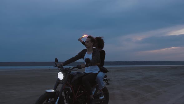 Young Couple Riding on Vintage Motorcycle with Red Burning Signal Fire After Sunset on Beach Slow