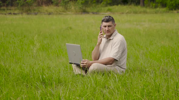 A Man with a Laptop Is Talking on the Phone on the Green Lawn