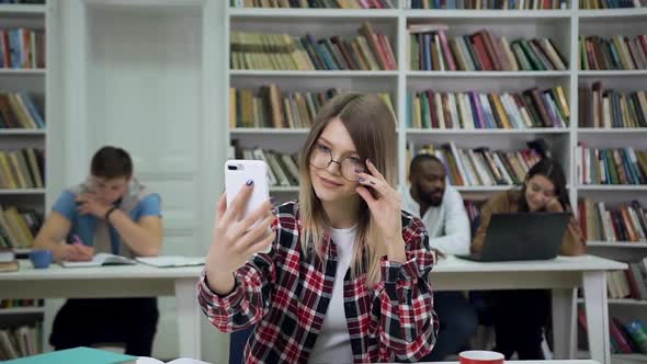 Female Student in Glasses Sitting in the Library and Watching Into the Phone Camera 