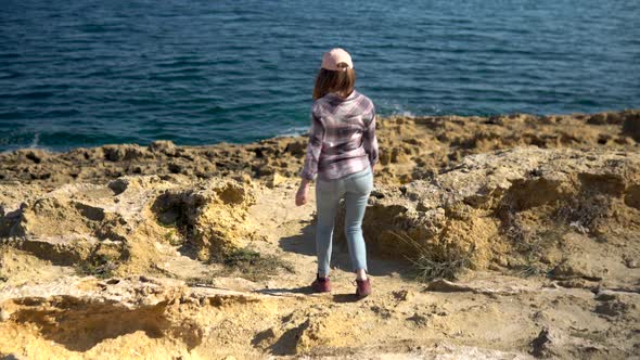 A Young Woman Sits Walking Toward the Sea on a Rock. The Girl Walks in Sunny Weather