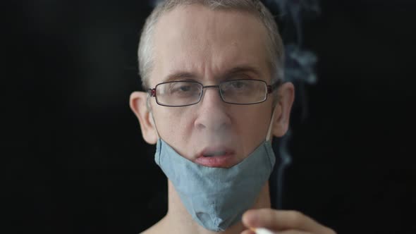 Caucasian Man Smoking a Cigarette in a Medical Mask on a Black Background