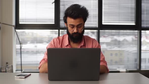 Front View of Thoughtful Bearded Young Business Man Typing on Laptop Computer Sitting at Desk in