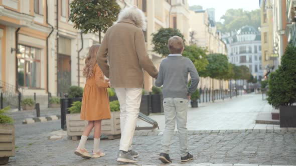 Wide Shot of Grandfather and Grandchildren Walking Along Paving Street on Sunny Day. Back View of