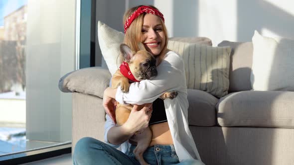 Young Woman in a White Shirt and Jeans Sits on the Floor Near the Sofa with Her Small French Bulldog