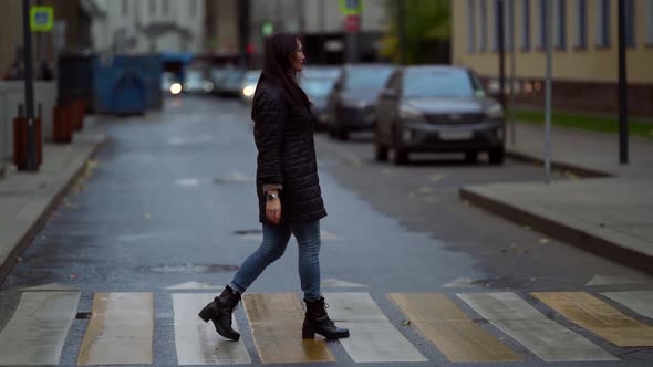 A Brunette with Long Hair Wearing a Knitted Sweater and a Dark Coat Walks Across a Pedestrian