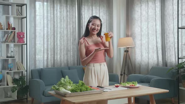 Asian Woman Holding And Pointing A Glass Of Orange Juice Before Smiling To Camera