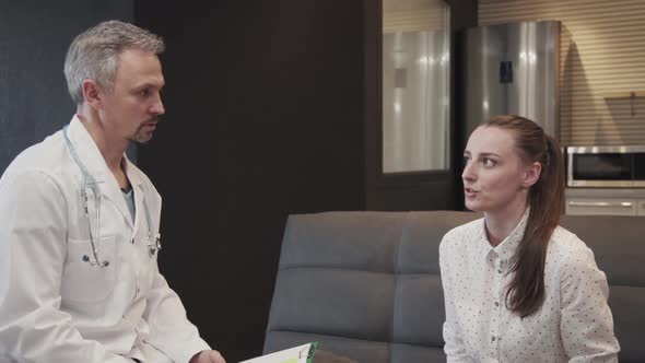 Male Doctor Talking To Female Patient and Writing While Sitting in Modern Clinic.