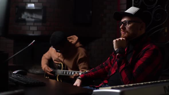 Professional Bearded Music Producer in Cap Sitting at Desk with Computer and Looking at Monitor