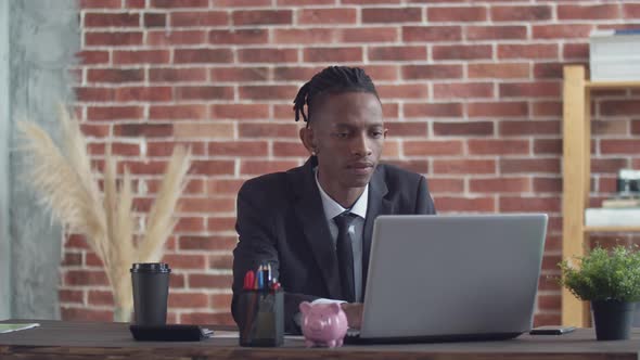 Black Male Businessman in a Strict Suit and with Serious Face Prints Text on a Laptop Sitting at