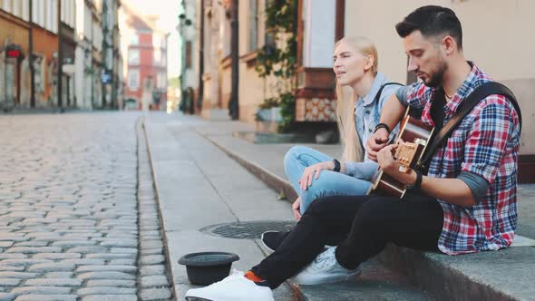 Young Man and Woman of Street Singers Sitting on Sidewalk, Playing Guitar and Singing