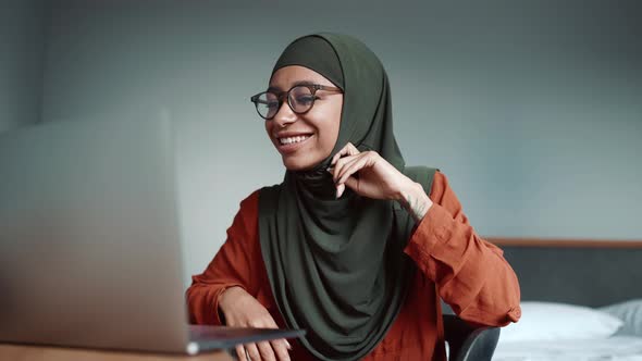 Muslim smiling woman wearing eyeglasses talking by video call on laptop