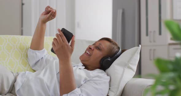 Happy african american senior woman wearing headphone lying on couch using smartphone and smiling