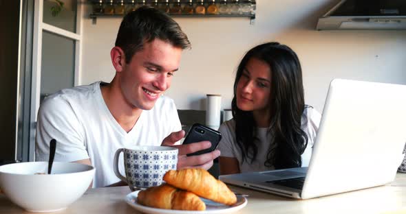 Couple using mobile phone and laptop while having breakfast