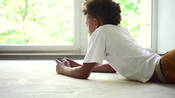 African Boy Playing on Smartphone Against Background of White Window with Summer Spbd Landscape