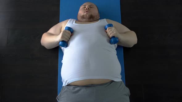 Purposeful Fat Man Lifting Dumbbells With Great Effort Lying on Mat, Top View