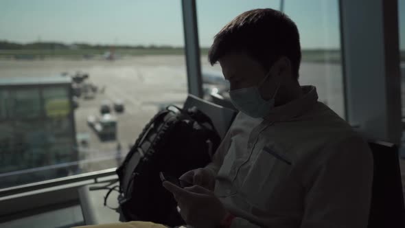 Man Passenger with Backpack in Protective Mask Waiting for Boarding a Plane Surfing Internet Using