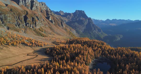 Backward Aerial Over Alpine Mountain Valley Lake and Orange Larch Forest Woods in Sunny Autumn