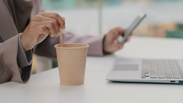 Closeup Female Hand of Sitting Businesswoman Stirring Coffee in Disposable Cup at Table with Laptop