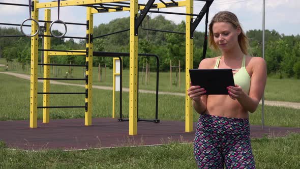 A Fit Beautiful Woman Works on a Tablet at an Outdoor Gym