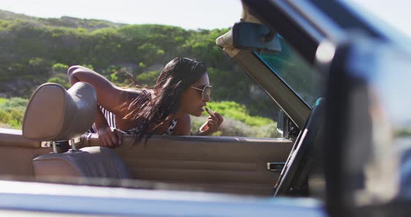 African american woman applying lipstick while looking in the mirror of convertible car on road