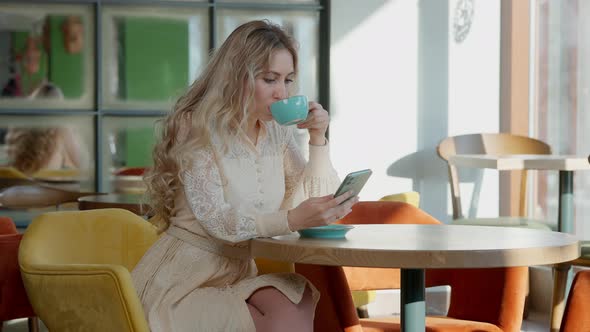 Slim Happy Relaxed Blond Woman Surfing Social Media on Smartphone Drinking Coffee in Restaurant