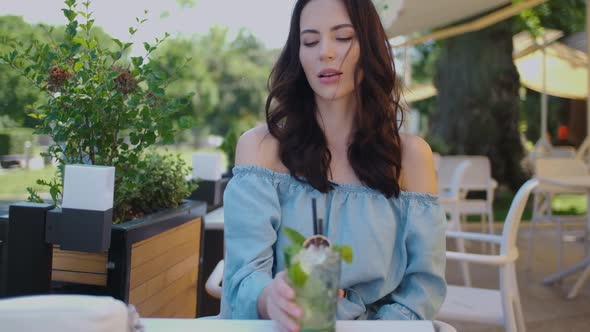 Beauiful Woman Sitting in Outdoor Restaurant and Sip Mojito Dring in Summer Hot Day