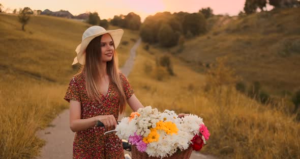 Young Smiling Blonde in Hat and Dress Walking in Dress with Bike and Flowers in Basket