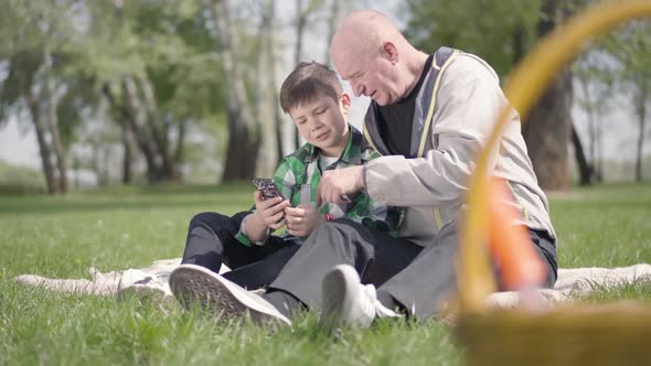 Portrait of Cute Senior Man Sitting with His Grandson on the Blanket in the Park