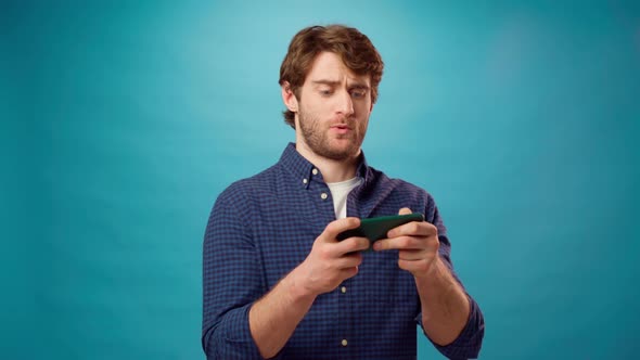 Young Man in Casual Shirt Playing Video Games on His Smartphone Against Blue Studio Background