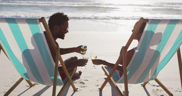 African american couple drinking wine together sitting on deck chairs at the beach