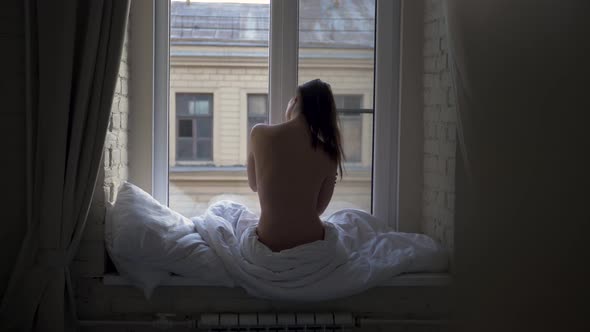 Woman Sits on the Window Topless Wrapped in a White Blanket