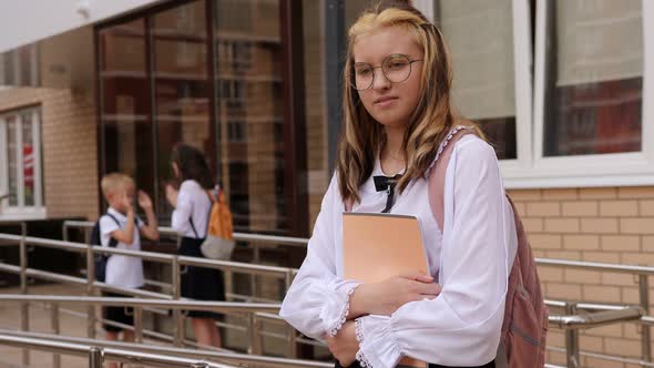 A Teenage Girl with Notebooks in the School Yard Near the Entrance to the School