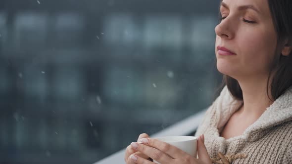 Caucasian Woman Stays on Balcony During Snowfall with Cup of Hot Coffee or Tea