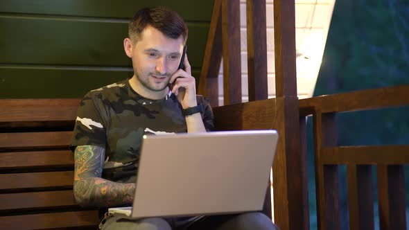 A Tattooed Man with a Laptop is Working on the Veranda of a Country House