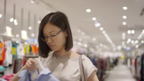 Asian Woman Choosing between Blouses in Shop and Posing for Camera