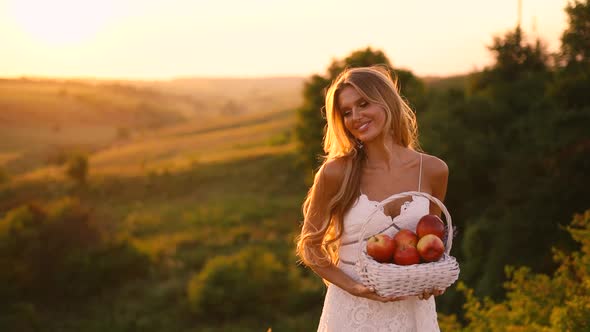 Beautiful sexy blonde girl in white dress posing in a field at sunset with a basket of fruit	