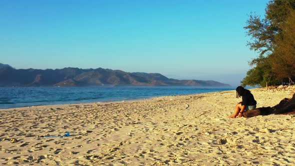 Young woman sitting on dried trunk playing with white sand of exotic beach, watching beautiful blue